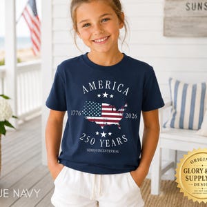 May include: A navy blue t-shirt featuring the text "AMERICA" above a map of the United States with the American flag design. The text "1776" and "2026" flank the map, with "250 YEARS" below. The shirt is worn by a young person.