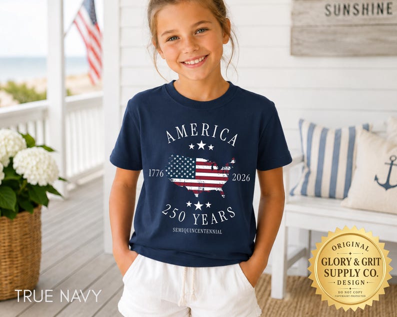 May include: A navy blue t-shirt featuring the text "AMERICA" above a map of the United States with the American flag design. The text "1776" and "2026" flank the map, with "250 YEARS" below. The shirt is worn by a young person.