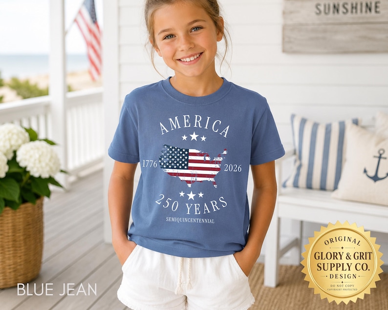 May include: A blue jean t-shirt with the word "AMERICA" above a map of the United States with the American flag design. The shirt also has the text "1776 2026 250 YEARS SEMIQUINCENTENNIAL". The shirt is worn by a young person.