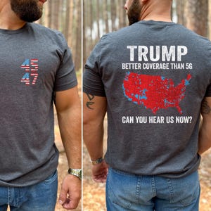 May include: A dark gray t-shirt with a red, white, and blue American flag design on the front and a map of the United States with red and blue markings on the back. The back of the shirt also has the text "TRUMP BETTER COVERAGE THAN 5G CAN YOU HEAR US NOW?"
