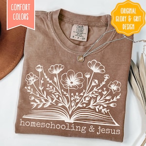 May include: A brown Comfort Colors t-shirt with a white floral design emerging from an open book, with the words "homeschooling & jesus" printed below. The shirt has a crew neck and short sleeves. A silver necklace with a pendant is on the shirt.