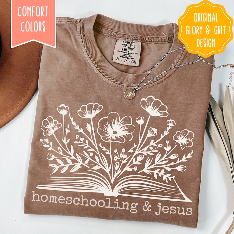 May include: A brown Comfort Colors t-shirt with a white floral design emerging from an open book, with the words "homeschooling & jesus" printed below. The shirt has a crew neck and short sleeves. A silver necklace with a pendant is on the shirt.