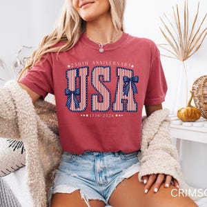 May include: Crimson-colored t-shirt with a patriotic "USA" design. The design features red and white stripes, blue bows, and the text "250th Anniversary 1776-2026". The shirt is a comfortable, casual style.