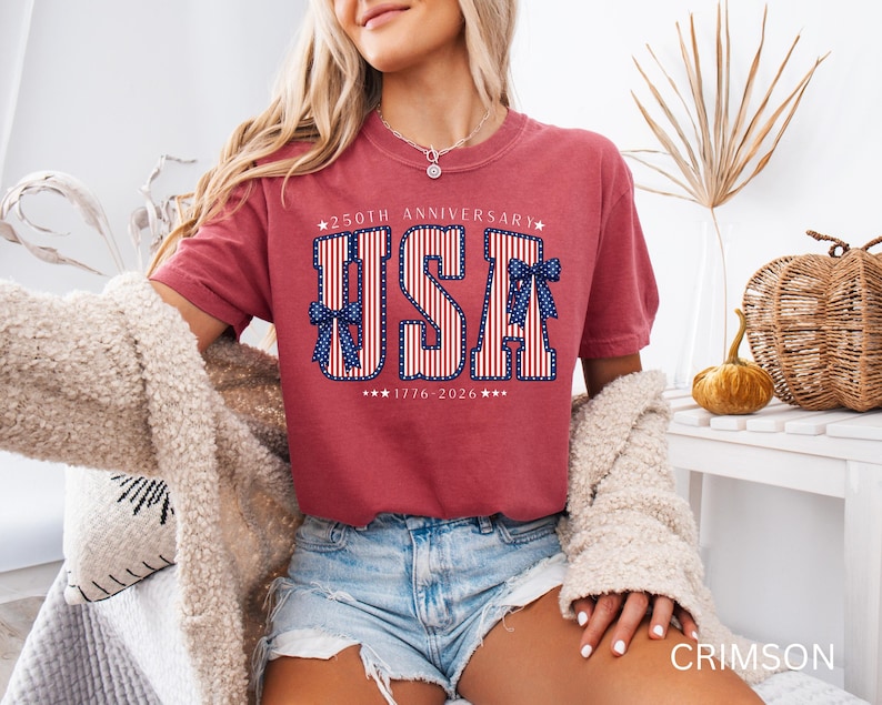 May include: Crimson-colored t-shirt with a patriotic "USA" design. The design features red and white stripes, blue bows, and the text "250th Anniversary 1776-2026". The shirt is a comfortable, casual style.