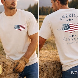 May include: Ivory-colored t-shirt featuring a distressed American flag design on the front pocket and back. The back design includes the text "AMERICA 250 YEARS SEMIQUINCENTENNIAL 1776-2026". The shirt is made of a soft material.