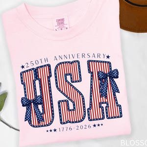May include: A light pink t-shirt with the word "USA" in red and white stripes, outlined in blue, and adorned with blue bows. The text "250th Anniversary 1776-2026" is also printed on the shirt. The shirt has a Comfort Colors tag.