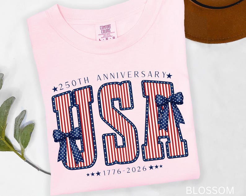 May include: A light pink t-shirt with the word "USA" in red and white stripes, outlined in blue, and adorned with blue bows. The text "250th Anniversary 1776-2026" is also printed on the shirt. The shirt has a Comfort Colors tag.