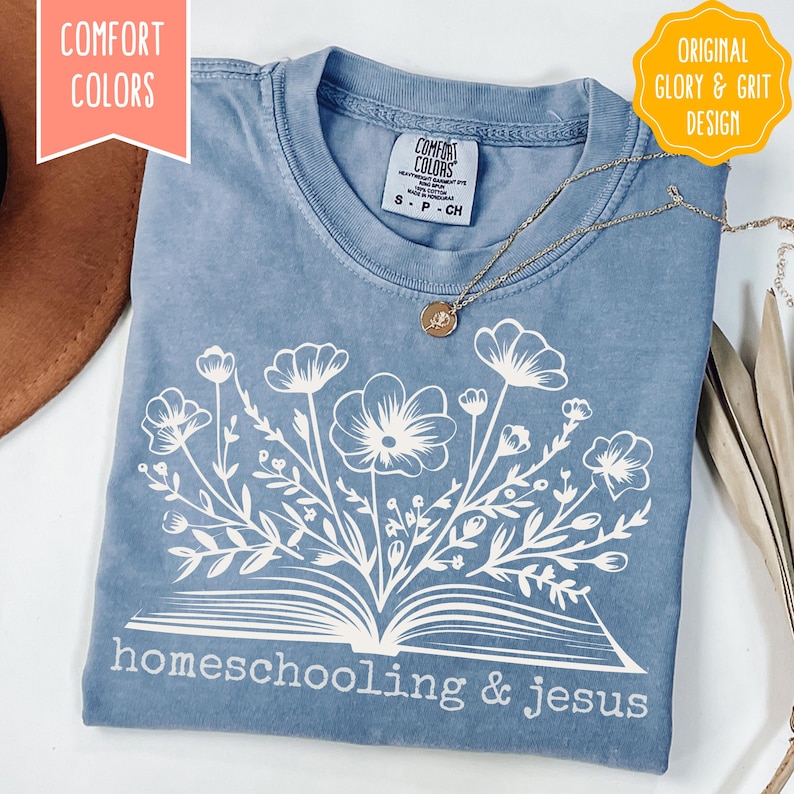 May include: A blue Comfort Colors t-shirt with a white floral design and the words "homeschooling & jesus." The shirt has a crew neck and short sleeves. A gold necklace with a pendant is draped across the shirt.