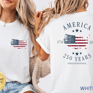 May include: White t-shirt featuring a distressed American flag design in the shape of the United States. The back of the shirt displays "AMERICA" above a flag graphic, with "1776" and "2026" flanking the flag, and "250 YEARS" below.