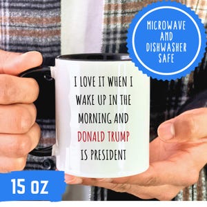 K&ouml;nnte beinhalten: Wei&szlig;er 444 ml Kaffeebecher mit schwarzem Henkel und Rand. Der Becher tr&auml;gt den Text "I LOVE IT WHEN I WAKE UP IN THE MORNING AND DONALD TRUMP IS PRESIDENT" in Schwarz und Rot. Ein blauer Kreis weist darauf hin, dass er mikrowellen- und sp&uuml;lmaschinenfest ist.