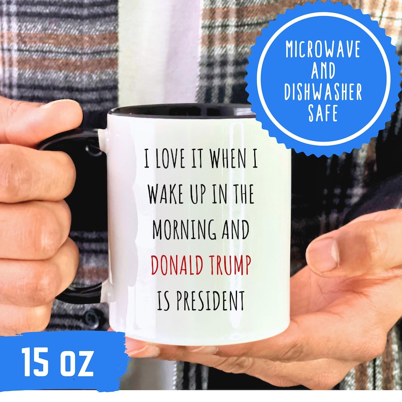K&ouml;nnte beinhalten: Wei&szlig;er 444 ml Kaffeebecher mit schwarzem Henkel und Rand. Der Becher tr&auml;gt den Text "I LOVE IT WHEN I WAKE UP IN THE MORNING AND DONALD TRUMP IS PRESIDENT" in Schwarz und Rot. Ein blauer Kreis weist darauf hin, dass er mikrowellen- und sp&uuml;lmaschinenfest ist.