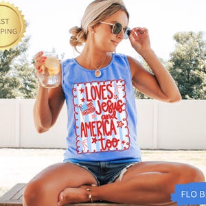 K&ouml;nnte beinhalten: Hellblaues Tanktop mit dem Text "Loves Jesus and America too" in Rot und Wei&szlig;. Das Design beinhaltet eine amerikanische Flagge und Sterne. Das Tanktop ist &auml;rmellos und hat einen runden Ausschnitt.