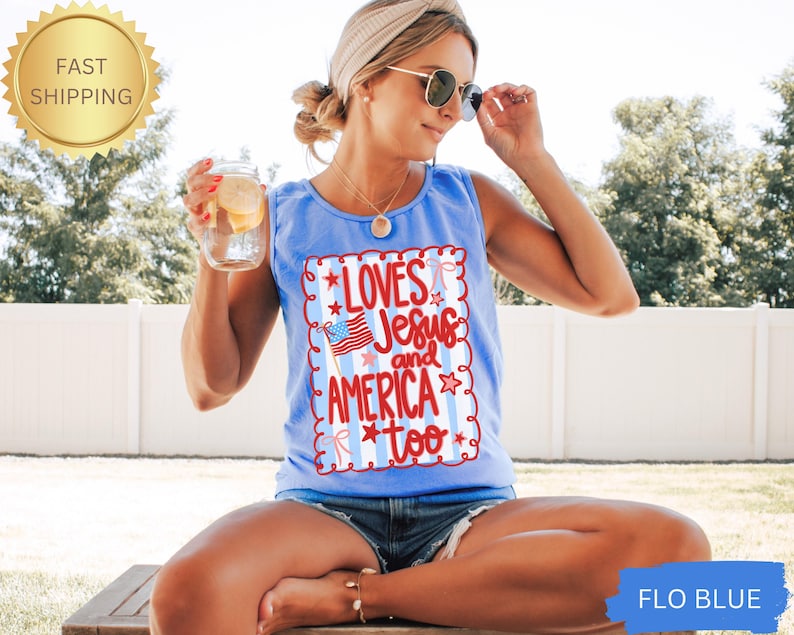 K&ouml;nnte beinhalten: Hellblaues Tanktop mit dem Text "Loves Jesus and America too" in Rot und Wei&szlig;. Das Design beinhaltet eine amerikanische Flagge und Sterne. Das Tanktop ist &auml;rmellos und hat einen runden Ausschnitt.