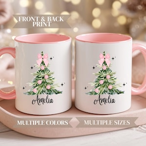 May include: Two white ceramic mugs with pink handles and interiors. Each mug features a Christmas tree design with a pink bow, ornaments, and the name "Amelia". The mugs are on a pink tray.