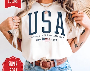 以下が含まれることがあります： アイボリー色のTシャツ。紺色のブロック体で「USA」と書かれています。その下に「UNITED STATES OF AMERICA EST 1776」の文字と小さなアメリカ国旗がプリントされています。丸首のTシャツです。