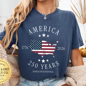 May include: A navy blue denim t-shirt with a graphic of the American flag in the shape of the United States. The shirt has the word "AMERICA" above the flag and "250 YEARS" below it. The years 1776 and 2026 are also printed on the shirt.