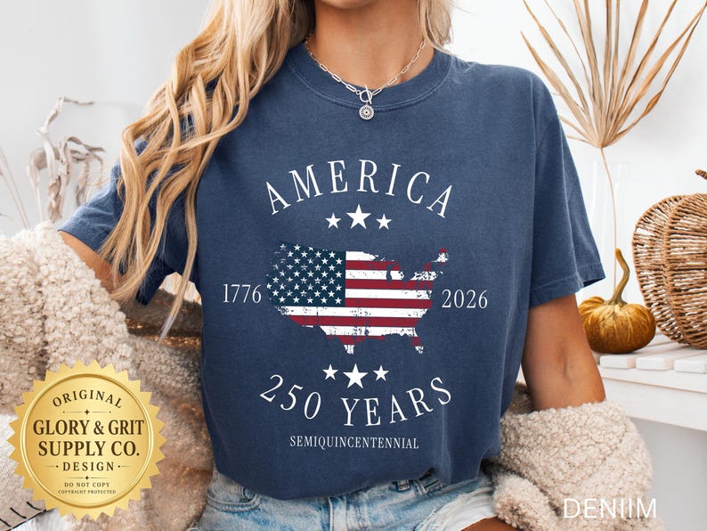 May include: A navy blue denim t-shirt with a graphic of the American flag in the shape of the United States. The shirt has the word "AMERICA" above the flag and "250 YEARS" below it. The years 1776 and 2026 are also printed on the shirt.