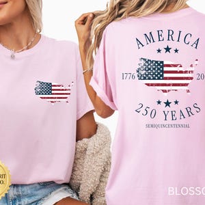May include: A light pink t-shirt with a distressed American flag design in the shape of the United States on the front and back. The back of the shirt also features the text "AMERICA 250 YEARS 1776-2026 SEMIQUINCENTENNIAL".