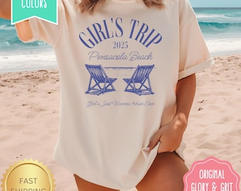 May include: Cream-colored t-shirt with the text "GIRL'S TRIP 2025 Pensacola Beach" in blue. The design includes two blue beach chairs and the phrase "Girl's Just Wanna Have Sun". The shirt has a relaxed fit.