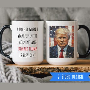 K&ouml;nnte beinhalten: Wei&szlig;e Keramik-Tasse mit schwarzem Griff und Rand. Eine Seite zeigt ein Portr&auml;t von Donald Trump vor dem Hintergrund der amerikanischen Flagge. Die andere Seite tr&auml;gt den Text: &bdquo;ICH LIEBE ES, WENN ICH MORGENS AUFWACHE UND DONALD TRUMP PR&Auml;SIDENT IST.&ldquo;
