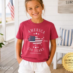 May include: A crimson t-shirt with the word "AMERICA" above a map of the United States with the American flag inside. The text "250 YEARS SEMIQUINCENTENNIAL" is below the map. The shirt is worn by a young person.