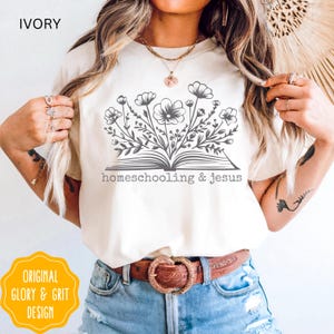 May include: Ivory-colored t-shirt featuring a floral design emerging from an open book, with the words "homeschooling & jesus" printed below. The shirt also has the text "ORIGINAL GLORY & GRIT DESIGN" in a yellow circle.