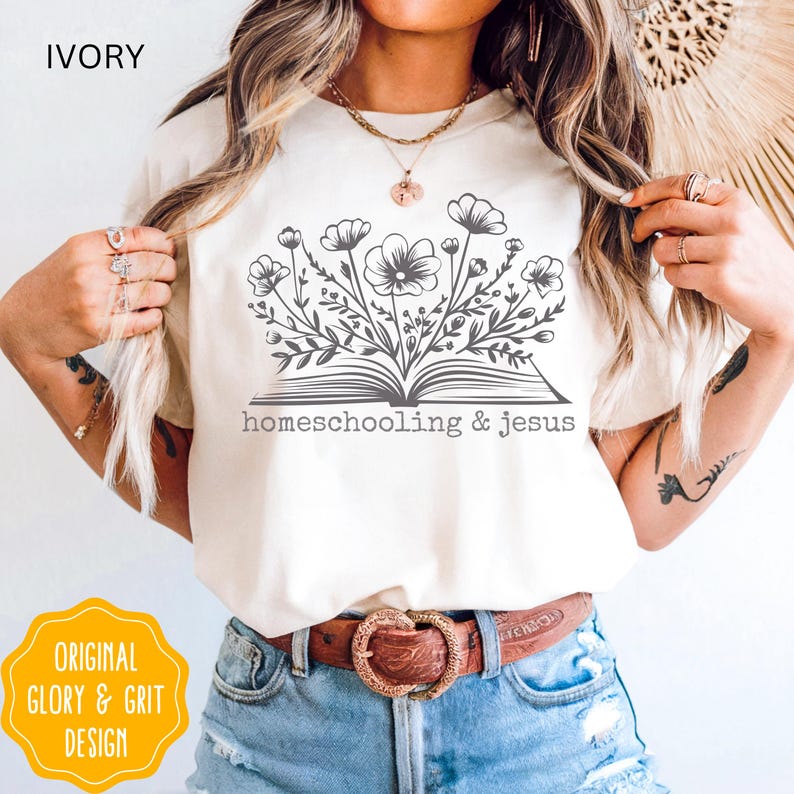 May include: Ivory-colored t-shirt featuring a floral design emerging from an open book, with the words "homeschooling & jesus" printed below. The shirt also has the text "ORIGINAL GLORY & GRIT DESIGN" in a yellow circle.