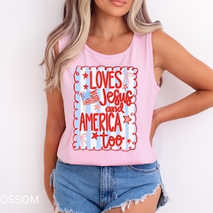 K&ouml;nnte beinhalten: Ein hellrosa Tanktop mit dem Schriftzug "LOVES Jesus and AMERICA too" in roter Schrift. Das Design beinhaltet eine amerikanische Flagge und ist von einem dekorativen Rahmen mit Sternen und Streifen umgeben.
