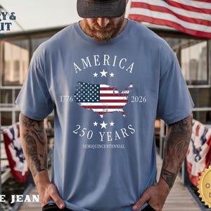 May include: A blue jean t-shirt with the text "AMERICA" and "250 YEARS" in white. The shirt features a graphic of the American flag within the shape of the United States, with the years 1776 and 2026. The shirt has a round neck.