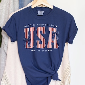 May include: Navy blue t-shirt with the word "USA" in red and white stripes, accented with blue bows and stars. The text "250th Anniversary" and "1776-2026" are also displayed. The shirt is hanging on a wooden hanger.