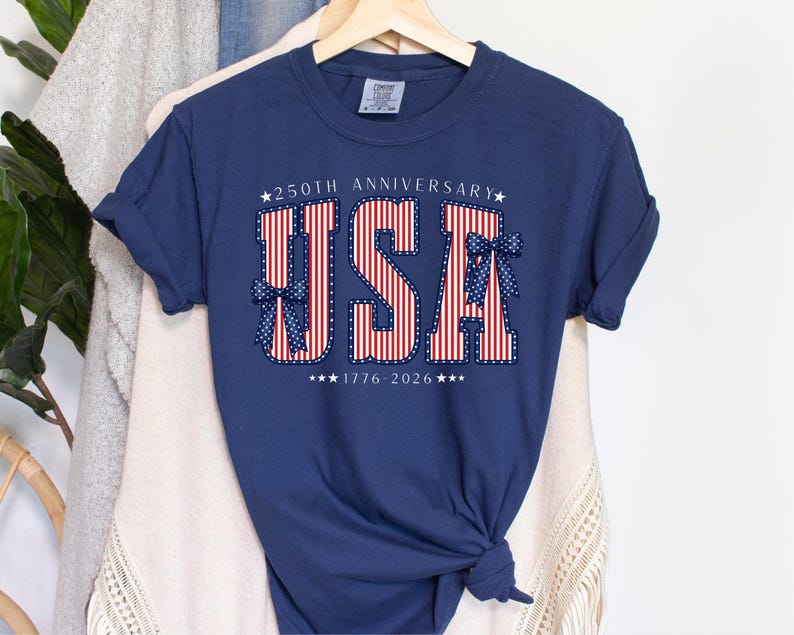 May include: Navy blue t-shirt with the word "USA" in red and white stripes, accented with blue bows and stars. The text "250th Anniversary" and "1776-2026" are also displayed. The shirt is hanging on a wooden hanger.