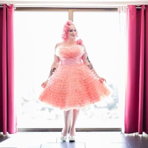 Puede incluir: Una mujer con el pelo rosa lleva un vestido de tul rosa con una falda con volantes. Está de pie frente a una ventana con cortinas rosas.