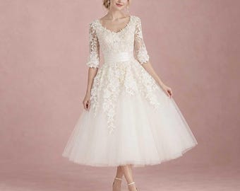 May include: A tea-length dress in ivory featuring a fitted bodice with three-quarter length sleeves, a defined waistline, and a full tulle skirt. The bodice and sleeves are adorned with intricate floral lace appliqués that extend down.
