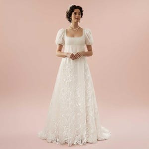 Puede incluir: Un vestido de novia blanco con escote cuadrado y mangas cortas abullonadas. El vestido presenta intrincados detalles de encaje floral en el corpiño y la falda. La modelo lleva un collar de perlas y una diadema, sobre un fondo rosa.