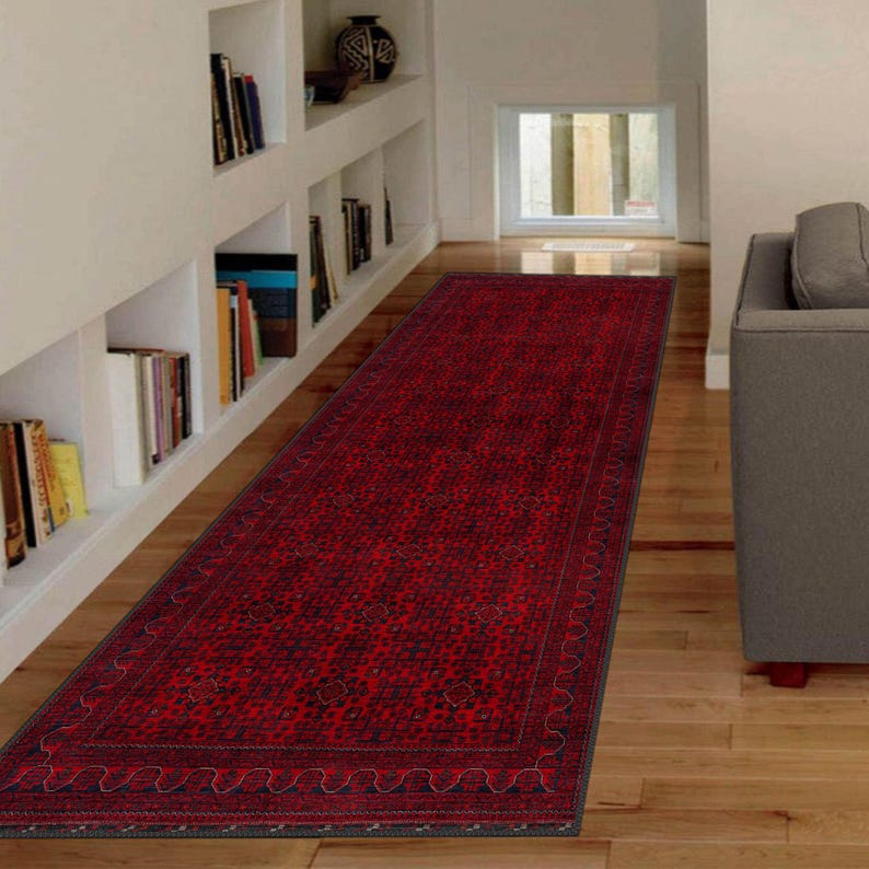 May include: A red runner rug with a traditional Persian design. The rug has a dark blue border with a repeating pattern. The rug is laid out on a hardwood floor in a hallway.