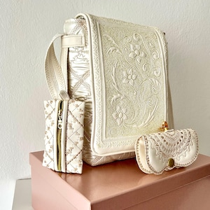 Könnte beinhalten: Elfenbeinfarbenes Handtaschen-Set mit einer Umhängetasche mit floraler Stickerei, einem kleinen Reißverschlussetui und einem Brillenetui. Das Set wird auf einer rosa Box präsentiert, die die feinen Details und Texturen der Accessoires zeigt.