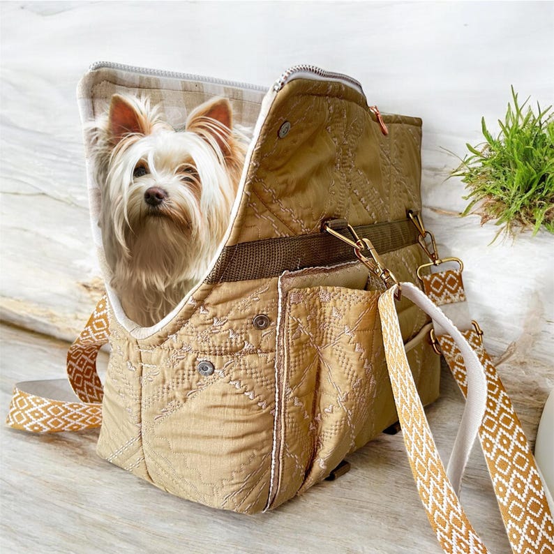 May include: Tan quilted pet carrier with a small dog inside. The carrier has a zippered top, multiple pockets, and a patterned shoulder strap. The dog has long, light-colored fur and is looking directly at the camera.