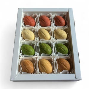 Peut inclure: Une boîte blanche contenant douze friandises ovales colorées. Les friandises sont disposées en trois rangées de quatre, avec des couleurs allant du rouge au jaune, en passant par le vert et le marron. La boîte a un intérieur blanc et est posée sur un fond blanc.
