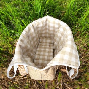 May include: A beige picnic basket with a gingham patterned interior and a matching cushion. The basket has white handles and is set on a bed of green grass.