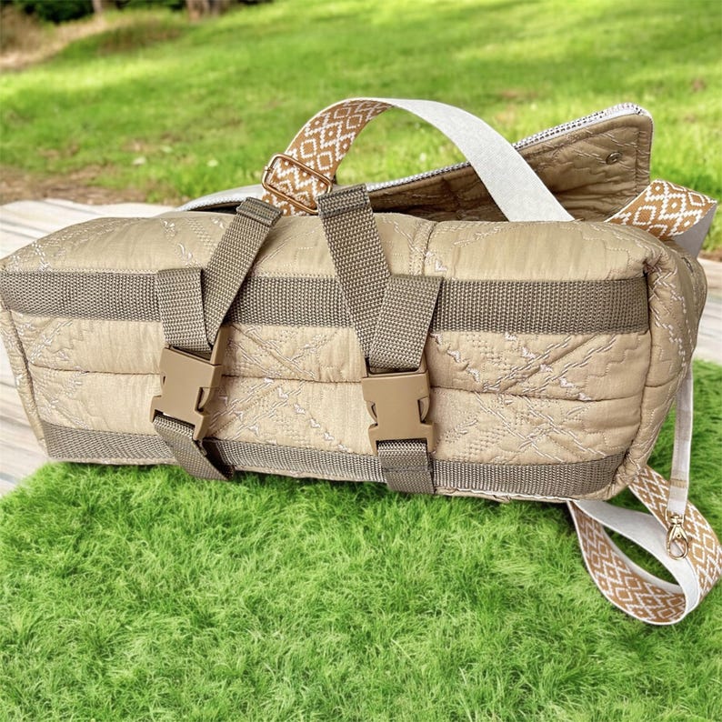 May include: A beige quilted duffel bag with brown straps and buckles. The bag has a patterned design on the straps and a white strap. The bag is sitting on a green surface.