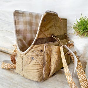 May include: A tan quilted fabric bag with a geometric pattern. The bag has a flap that opens to reveal a plaid interior. It features gold-colored hardware, a brown strap, and a patterned shoulder strap.
