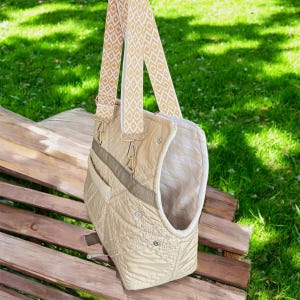 May include: A beige quilted pet carrier bag with a patterned strap. The bag has a rectangular shape with a curved opening and a small pocket on the side. The strap is beige with a white diamond pattern. The bag is hanging on a wooden bench.