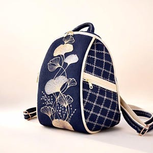 Könnte beinhalten: Marineblauer Rucksack mit floraler Stickerei. Er hat eine cremefarbene Paspel und einen Reißverschluss sowie ein Steppmuster an der Seite. Die Träger sind marineblau mit cremefarbenen Akzenten. Höhe ca. 30 cm.