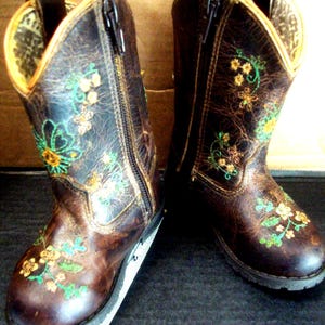 May include: A pair of brown leather cowboy boots with embroidered floral designs in yellow, green, and blue. The boots have zippers and a yellow lining. The boots are designed for children.