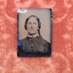 May include: A vintage portrait of a woman in a dark dress with a white collar, set within a rectangular frame. The frame is surrounded by a plush, peach-colored fabric with ornate designs, and a dark blue border with gold detailing.
