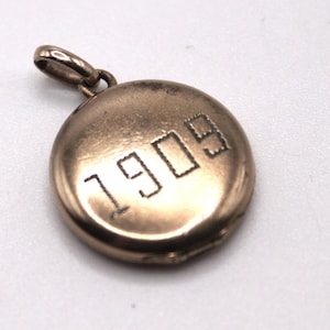 May include: A vintage gold-coloured pendant with a circular design. The pendant features the engraved year "1909" in a block font. It has a small loop at the top for attaching to a chain or bracelet. The pendant is likely made of metal.