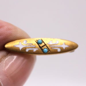 May include: A gold-toned bar pin with white enamel accents and two small turquoise-colored stones. The pin has an oval shape with a decorative design. The pin is held between two fingers.