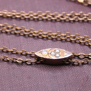 May include: Close-up of a gold-toned necklace with a decorative oval pendant. The pendant features small, round, and oval-shaped gemstones. The chain is made of small, interlocking links. The necklace rests on a purple fabric surface.