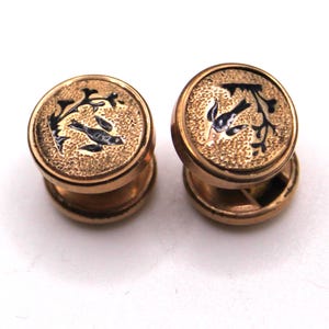 May include: A pair of gold-coloured cufflinks featuring a bird and floral motif. Each cufflink has a circular face with a detailed black and gold pattern. The design is consistent across both cufflinks, forming a matching set.
