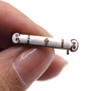 May include: A small, antique bar pin with a white enamel center and gold-toned accents. The pin features decorative scrollwork at each end and a small, diamond-shaped detail in the center. The pin is held in a hand against a white background.